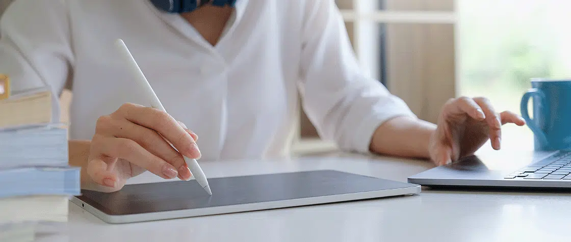 A person sketches on a drawing tablet with a white stylus in their right hand. In their left hand they're typing on the laptop keyboard that's next to them.