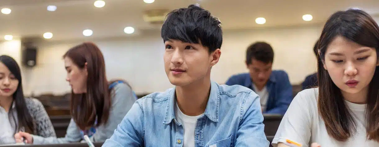 A man looks up and towards the speaker in a busy lecture hall. Around him are other students engrossed in their assignments or side conversations, the man is attentively paying attention to the speaker's presentation.