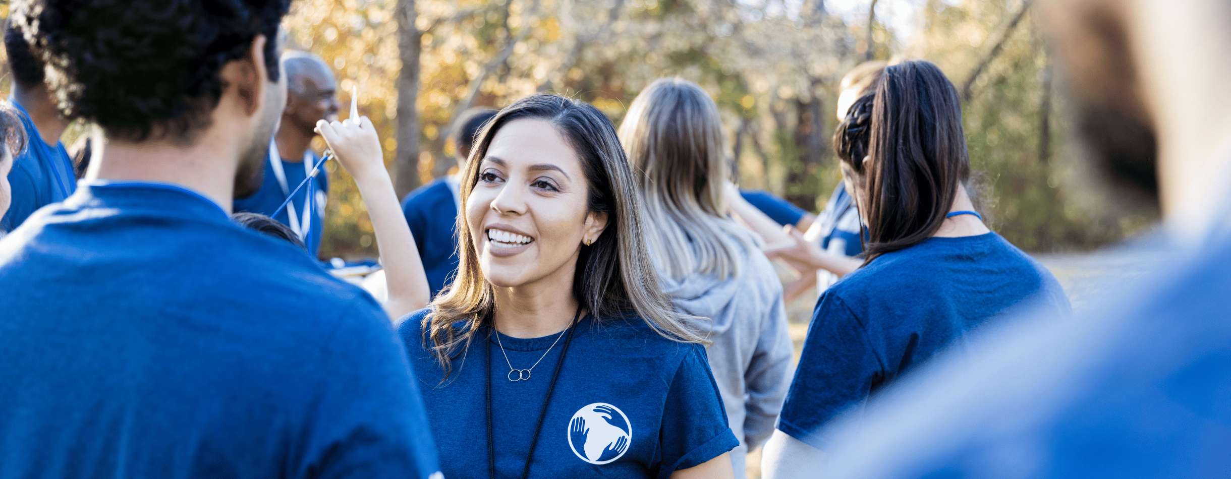 The photo centers on a woman smiling and talking to her acquaintance at a volunteer event. Both of them, along with the several other people in the background of the photo, are wearing navy colored shirts with a logo that designates them as part of the same organization and volunteer efforts. It is a small moment in a larger crowd of volunteers.