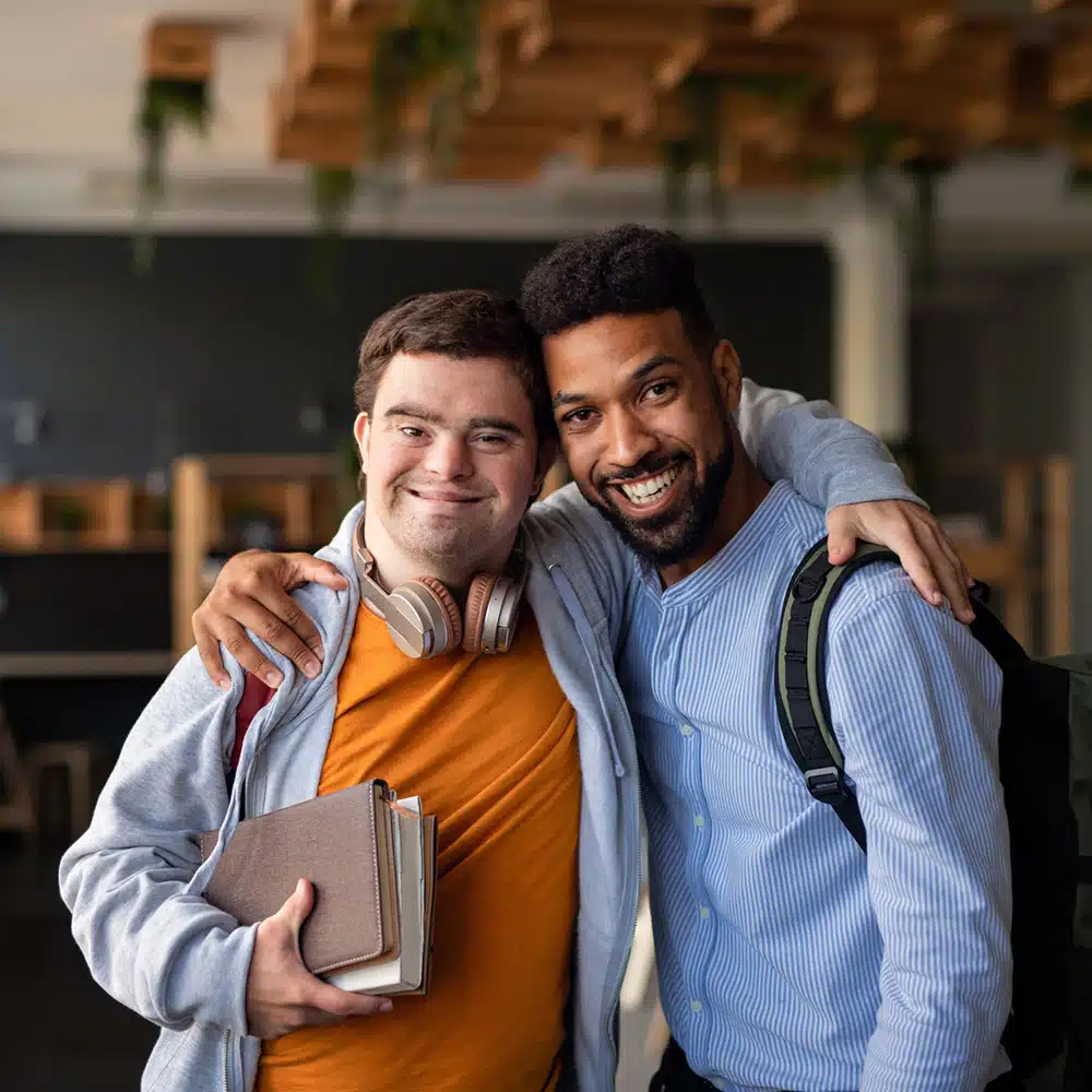 Young Man With Down Syndrome And His Tutor With Arms Around Looking At Camera Indoors At School