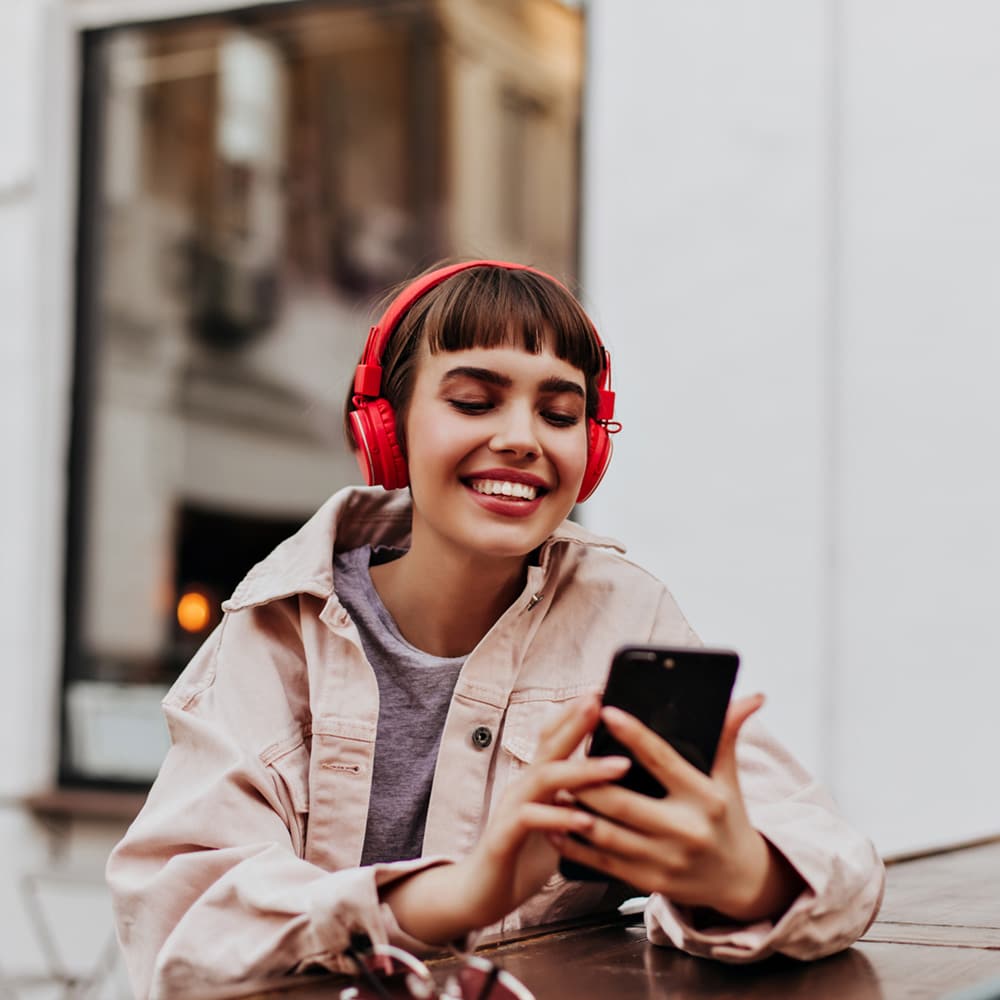 Positive Short Haired Girl In Red Headphones Smiles Outdoors. Brunette Woman In Beige Jacket Holding Phone And Listening To Music Outside.