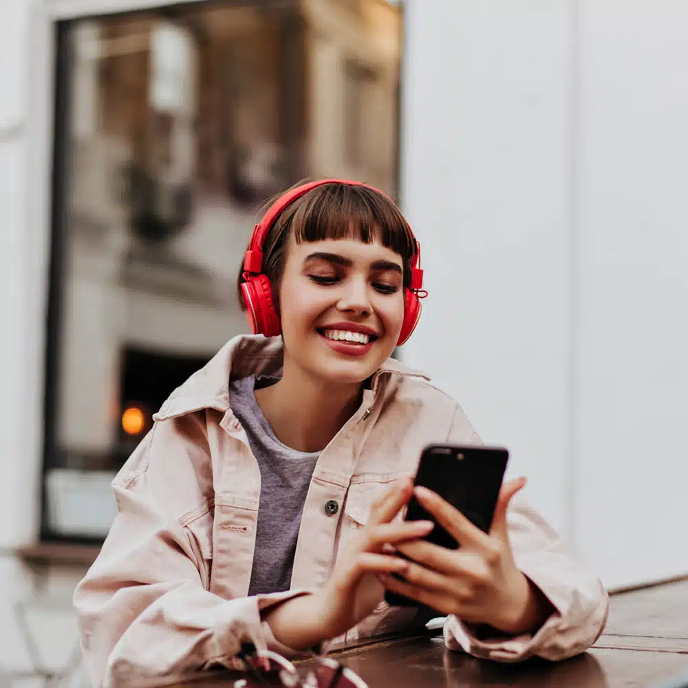 Positive Short Haired Girl In Red Headphones Smiles Outdoors. Brunette Woman In Beige Jacket Holding Phone And Listening To Music Outside.